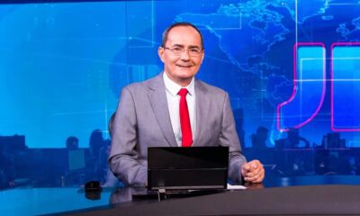Ayres Rocha sorri em estúdio de TV, terno cinza, fundo azul com mapa e logo JN