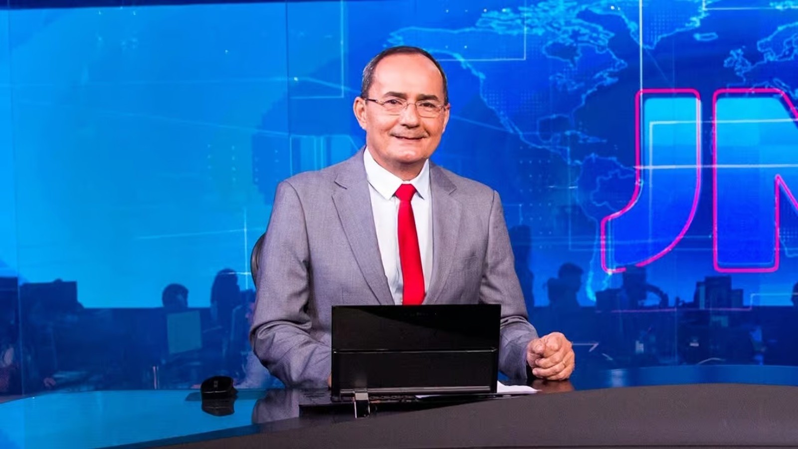 Ayres Rocha sorri em estúdio de TV, terno cinza, fundo azul com mapa e logo JN