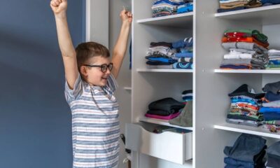 Menino sorrindo com braços erguidos e óculos ao lado de guarda-roupa infantil com roupas organizadas.