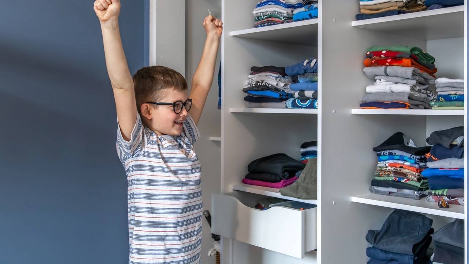Menino sorrindo com braços erguidos e óculos ao lado de guarda-roupa infantil com roupas organizadas.