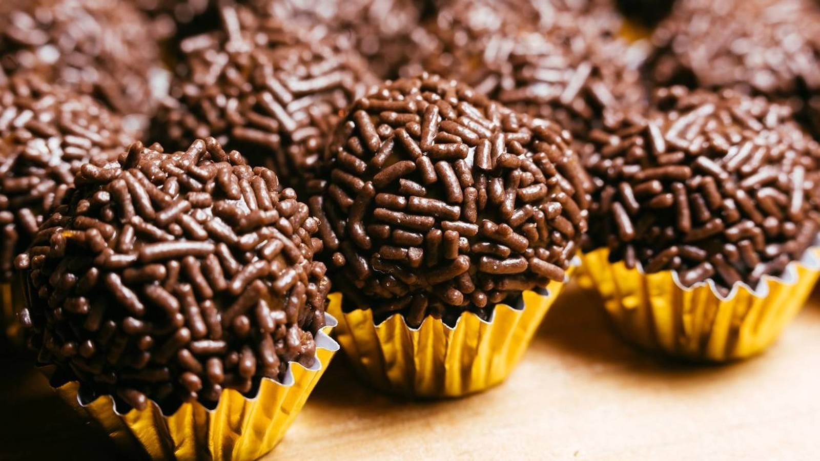 Vários brigadeiros de chocolate enrolados, com granulado e em forminhas douradas, em destaque