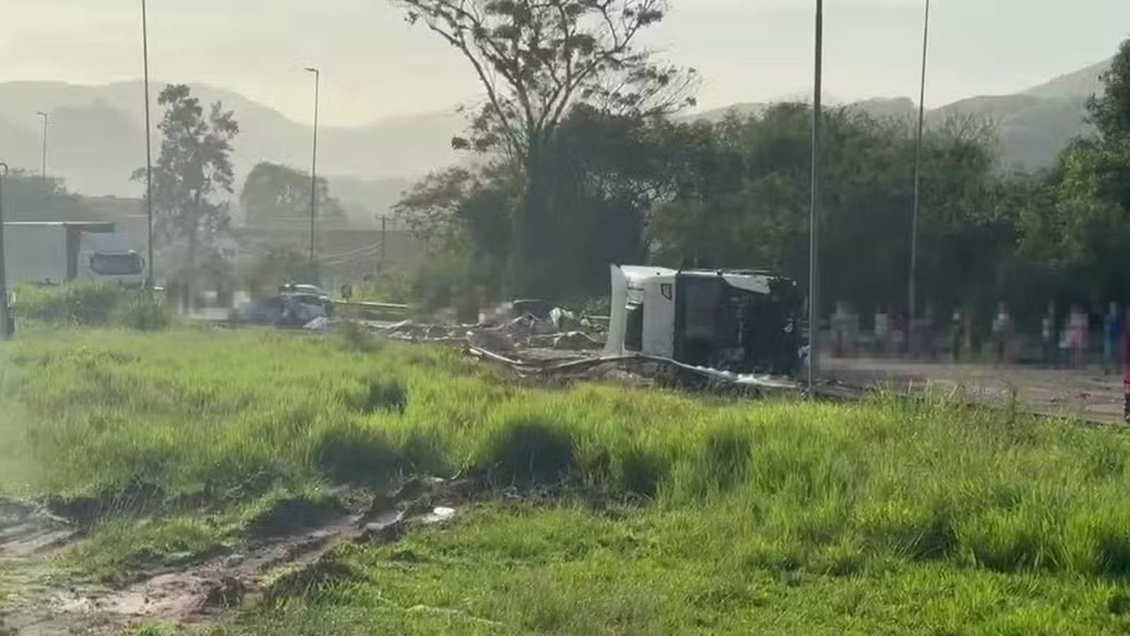Caminhão branco tombado em acostamento de rodovia, com detritos e grama ao redor