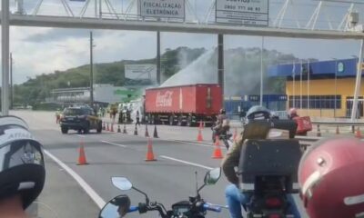 Caminhão Coca-Cola vermelho com água sendo jorrada, fumaça, cones, viatura PRF e prédio policial