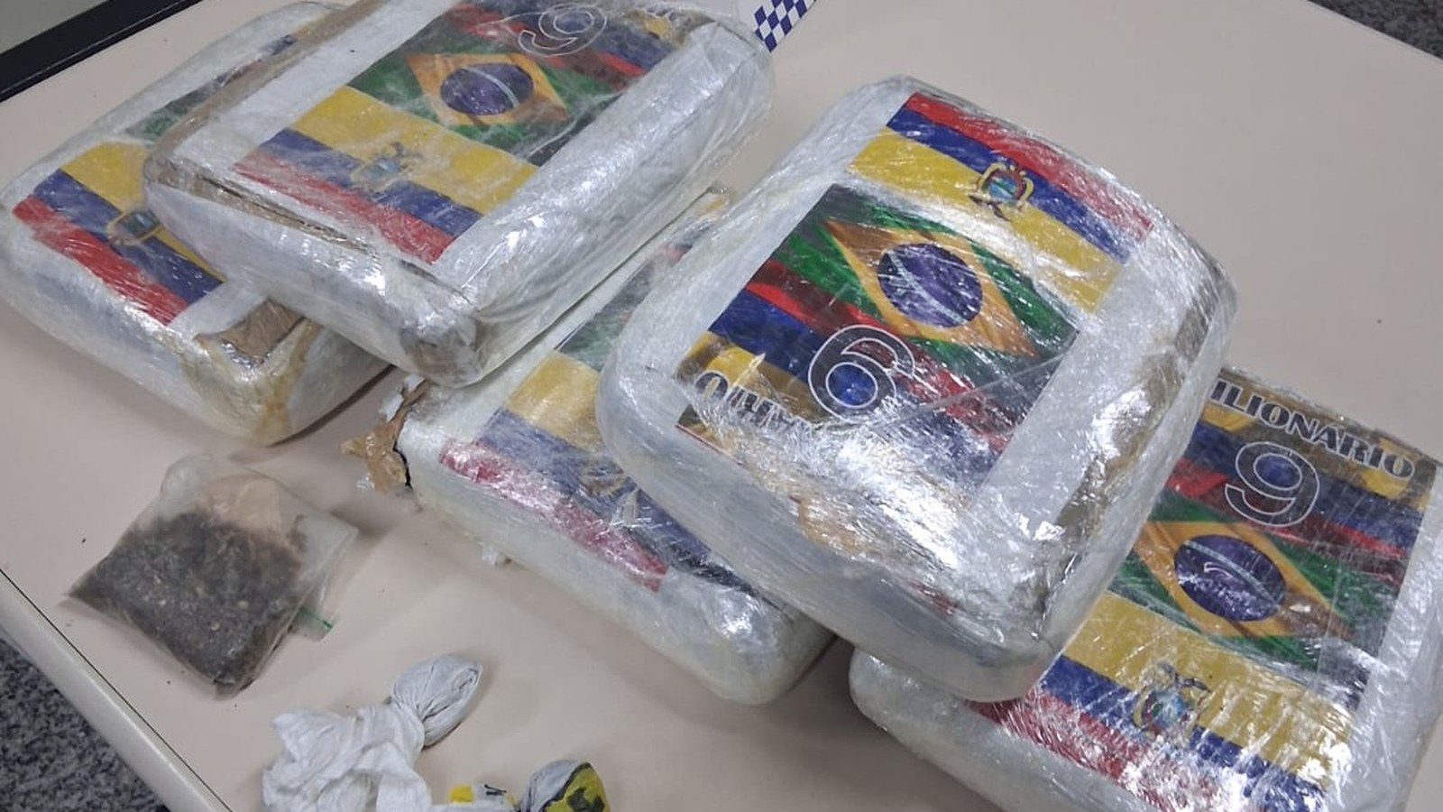 Blocos de maconha prensada com adesivos de bandeiras e 'MILIONARIO 9', e um saco pequeno de erva