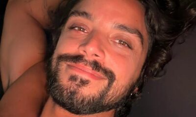 Homem com cabelo escuro cacheado, barba e bigode olhando e sorrindo para a câmera