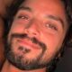 Homem com cabelo escuro cacheado, barba e bigode olhando e sorrindo para a câmera