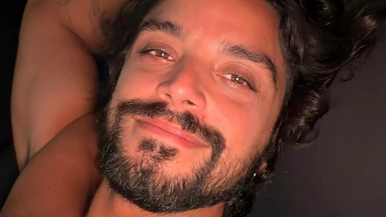 Homem com cabelo escuro cacheado, barba e bigode olhando e sorrindo para a câmera