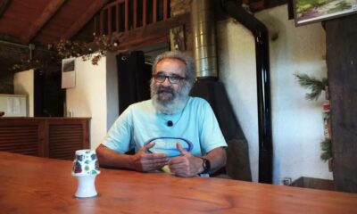 Homem barbudo de óculos e camiseta azul gesticula à mesa de madeira, com microfone de lapela