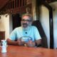 Homem barbudo de óculos e camiseta azul gesticula à mesa de madeira, com microfone de lapela