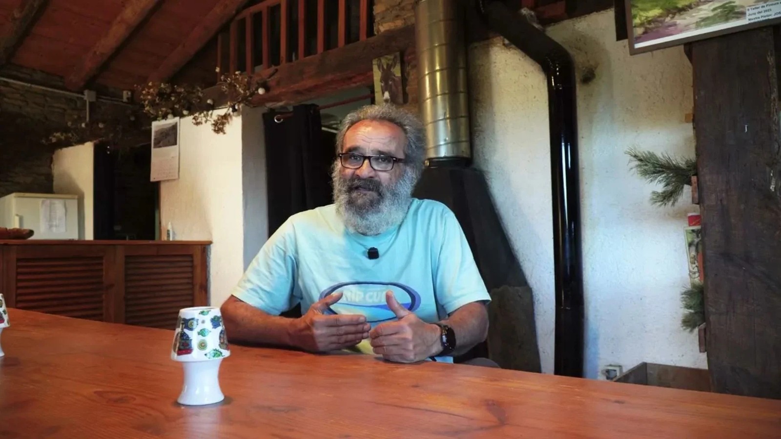 Homem barbudo de óculos e camiseta azul gesticula à mesa de madeira, com microfone de lapela