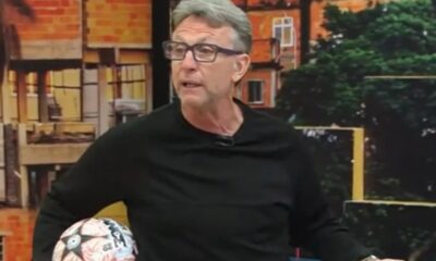 Craque Neto, homem de óculos e camisa preta, segura bola de futebol em estúdio