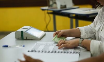 Mãos de mesária com documento eleitoral e lista de votação; urna eletrônica ao fundo