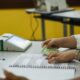 Mãos de mesária com documento eleitoral e lista de votação; urna eletrônica ao fundo