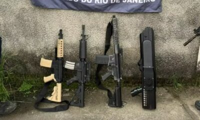 Três fuzis e um dispositivo antidrone apreendidos contra muro, banner policial atrás