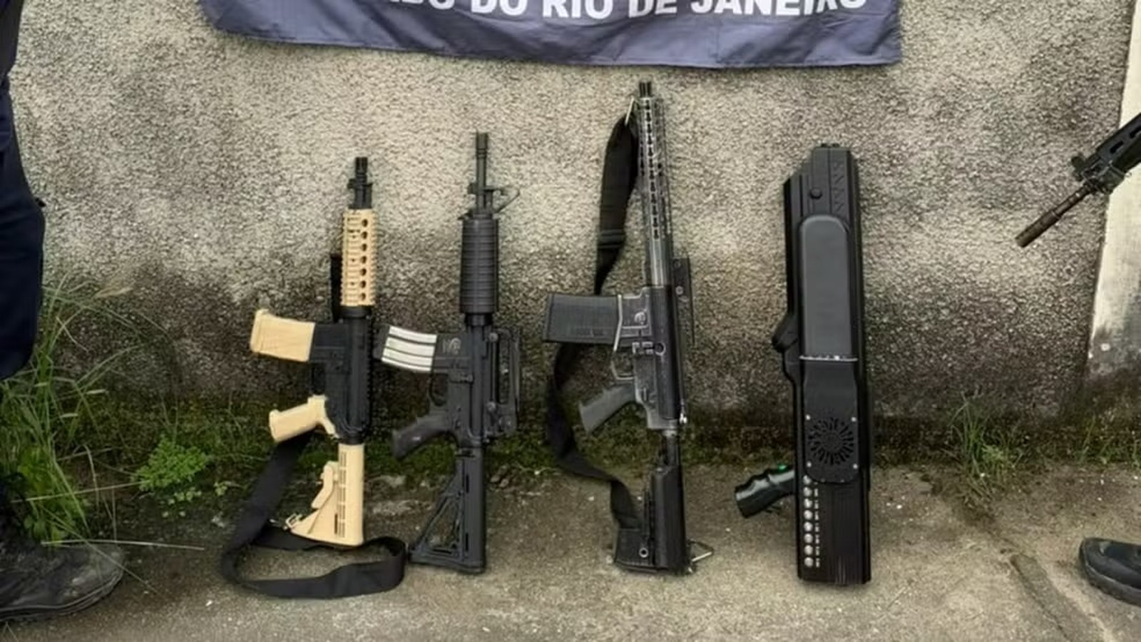 Três fuzis e um dispositivo antidrone apreendidos contra muro, banner policial atrás