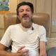 Youtuber Lito Sousa, com barba e camiseta clara, sentado em poltrona, com microfone e soro intravenoso