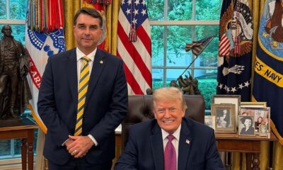 Flávio Bolsonaro e Donald Trump sorriem no Salão Oval, com bandeiras e estátua ao fundo