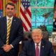Flávio Bolsonaro e Donald Trump sorriem no Salão Oval, com bandeiras e estátua ao fundo