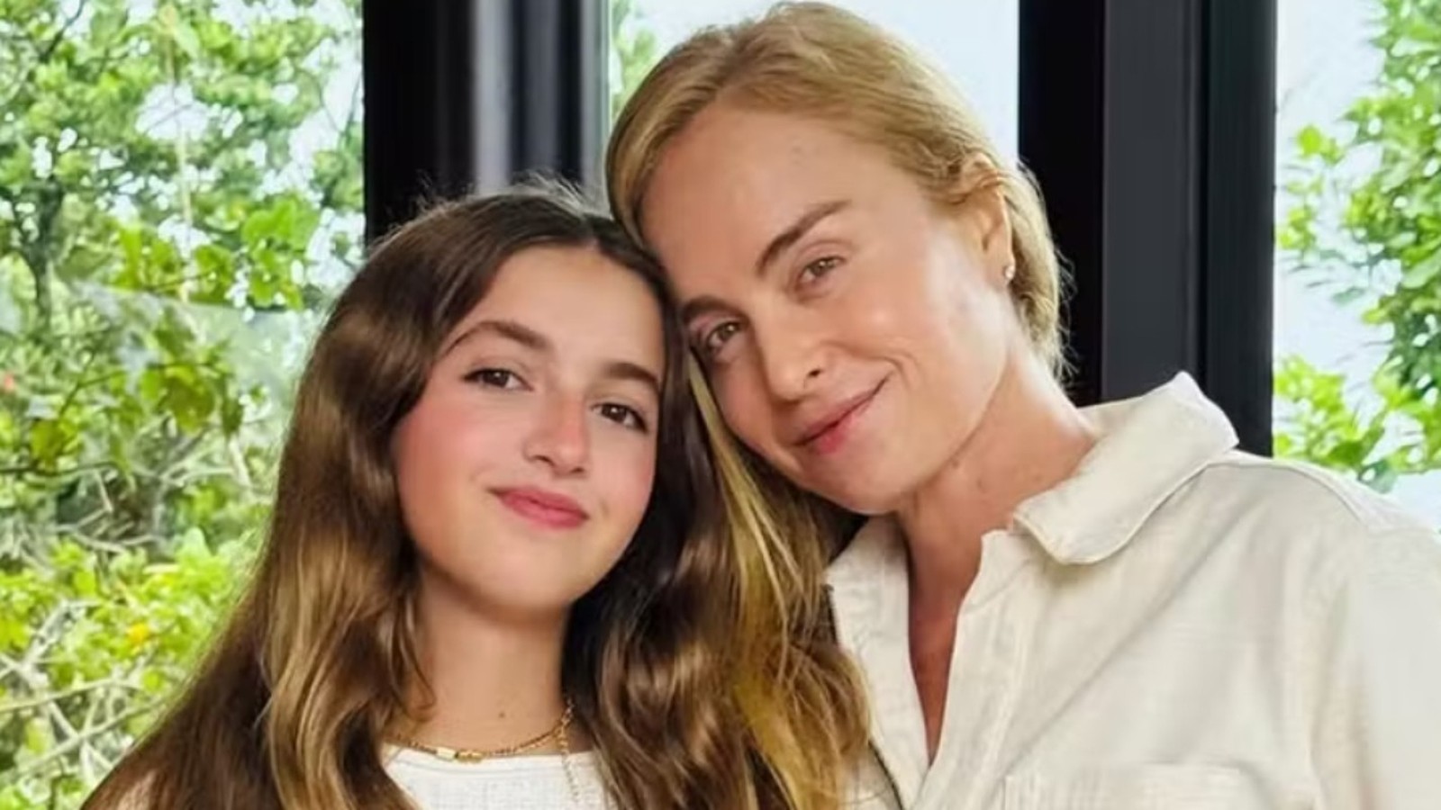 Angélica e sua filha Eva, sorrindo e com cabeças juntas, em frente a uma folhagem verde