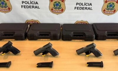 Quatro pistolas pretas e carregadores sobre mesa de madeira com caixas e banner da Polícia Federal.