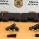 Quatro pistolas pretas e carregadores sobre mesa de madeira com caixas e banner da Polícia Federal.