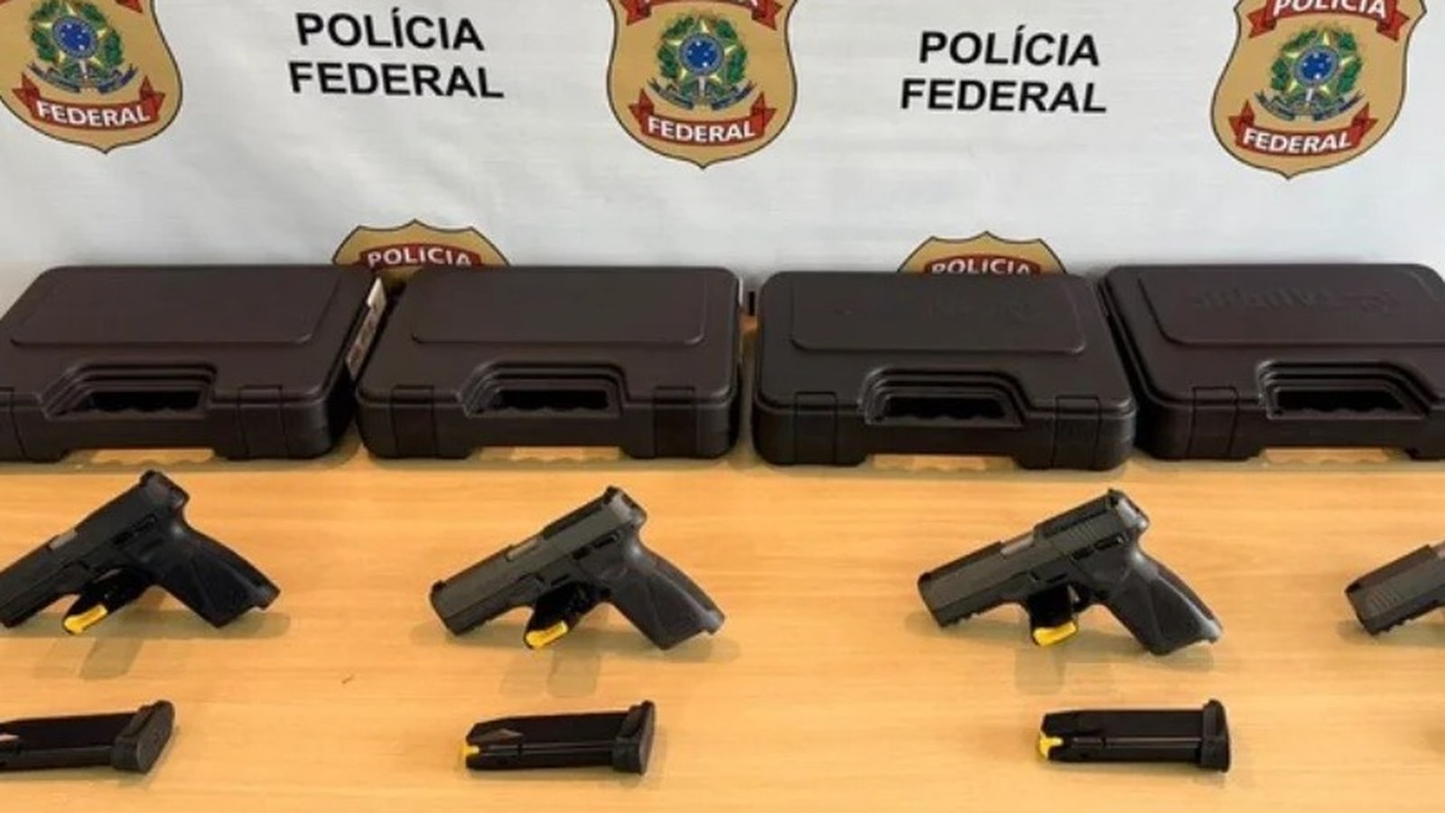 Quatro pistolas pretas e carregadores sobre mesa de madeira com caixas e banner da Polícia Federal.