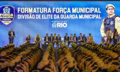 Formatura da Força Municipal com agentes uniformizados, autoridades no palco e telão ao fundo