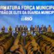 Formatura da Força Municipal com agentes uniformizados, autoridades no palco e telão ao fundo