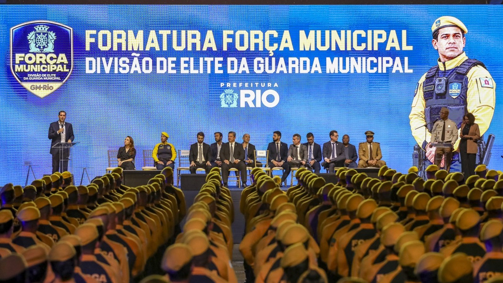 Formatura da Força Municipal com agentes uniformizados, autoridades no palco e telão ao fundo