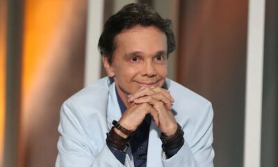 Cantor Netinho, com blazer azul claro e pulseiras de couro, sorri com as mãos unidas