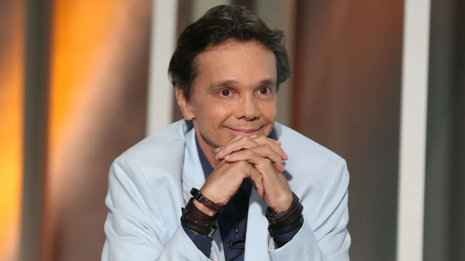 Cantor Netinho, com blazer azul claro e pulseiras de couro, sorri com as mãos unidas