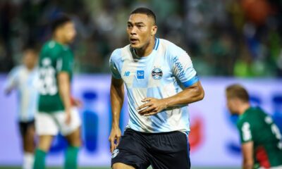 Jogador Carlos Vinícius em campo com camisa listrada azul e branca do Grêmio e shorts pretos
