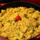 Panela de ferro vermelha com farofa amarela, salpicada com cheiro-verde e uma pimenta vermelha