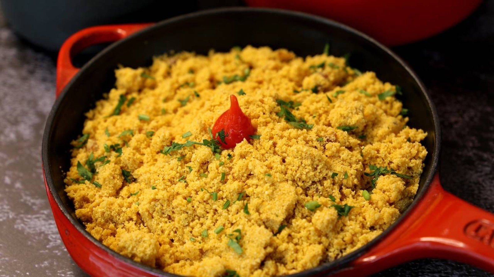 Panela de ferro vermelha com farofa amarela, salpicada com cheiro-verde e uma pimenta vermelha