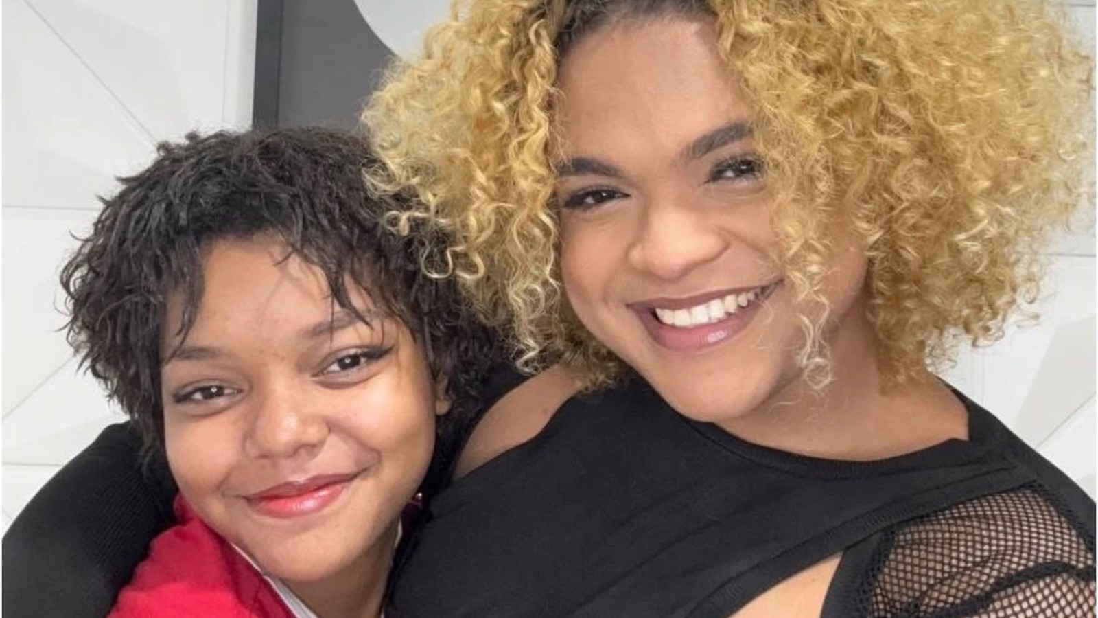 Duas jovens sorridentes, uma com cabelo crespo escuro, outra com cabelo crespo loiro, se abraçando