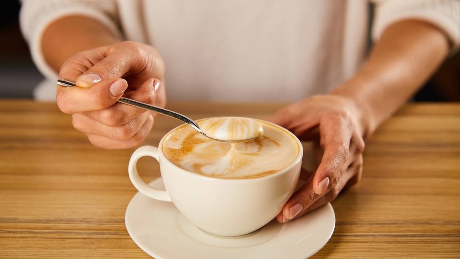 Mãos seguram colher com espuma de café cremoso em xícara branca sobre pires