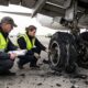 Dois investigadores examinam pneus estourados de avião no asfalto, um com colete "INVESTIGATOR" visível