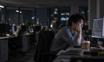 Homem asiático cansado olha monitores de computador em escritório noturno com luzes da cidade ao fundo