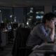 Homem asiático cansado olha monitores de computador em escritório noturno com luzes da cidade ao fundo
