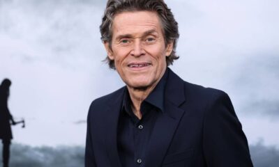 Retrato de Willem Dafoe sorrindo, vestindo terno escuro, com fundo cinza desfocado e silhueta distante