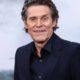 Retrato de Willem Dafoe sorrindo, vestindo terno escuro, com fundo cinza desfocado e silhueta distante