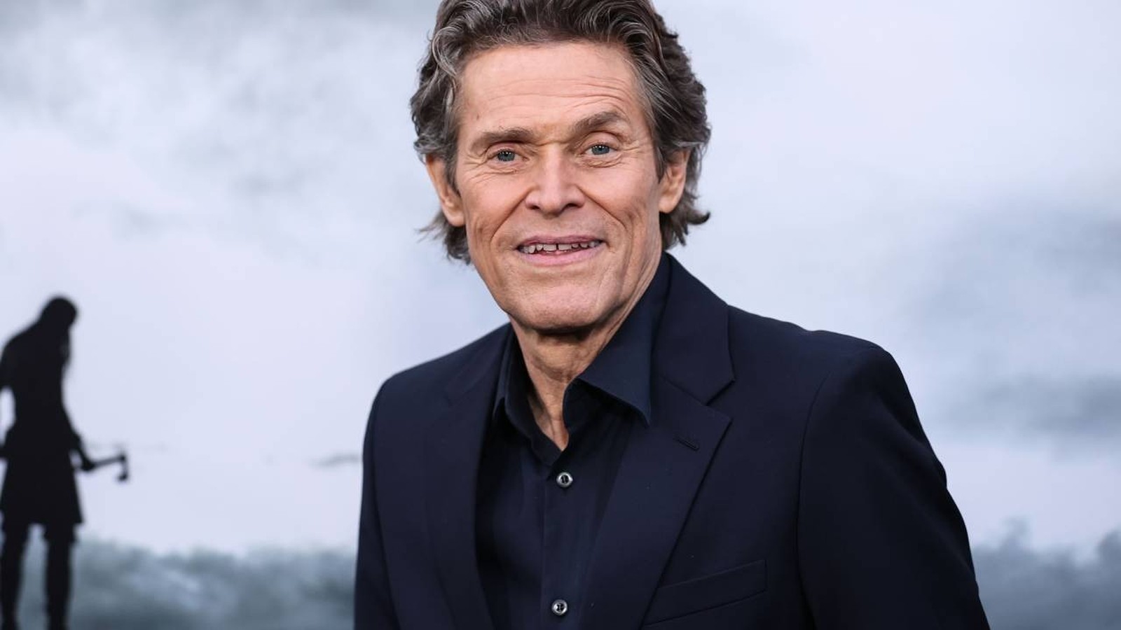 Retrato de Willem Dafoe sorrindo, vestindo terno escuro, com fundo cinza desfocado e silhueta distante