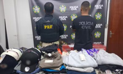 Dois policiais de costas, um da PRF, exibem roupas de marca apreendidas sobre mesa