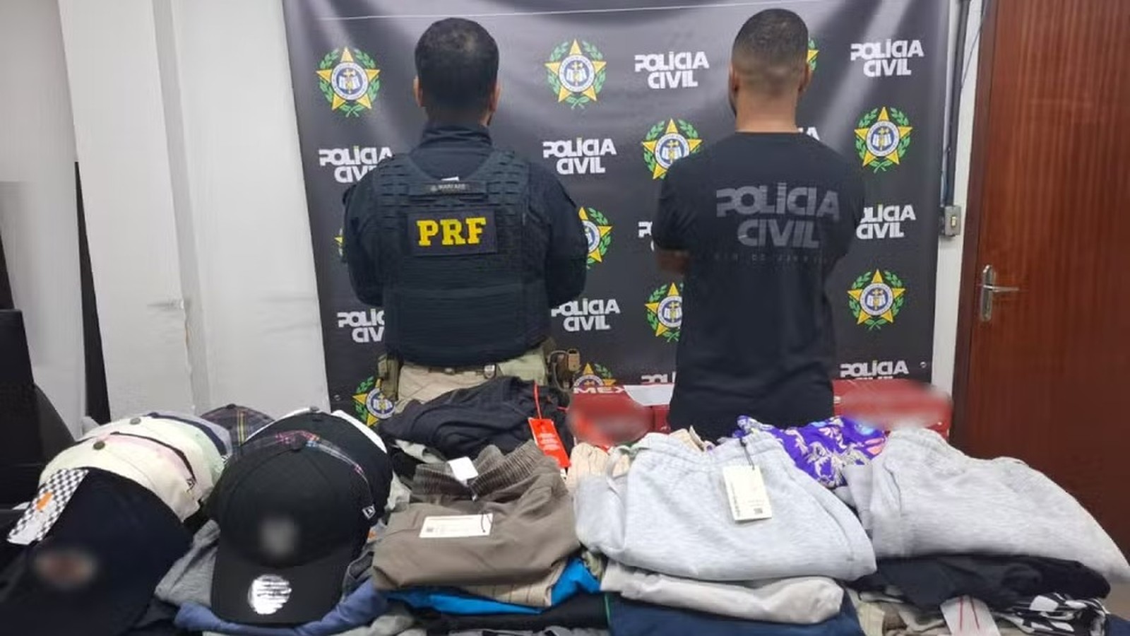Dois policiais de costas, um da PRF, exibem roupas de marca apreendidas sobre mesa