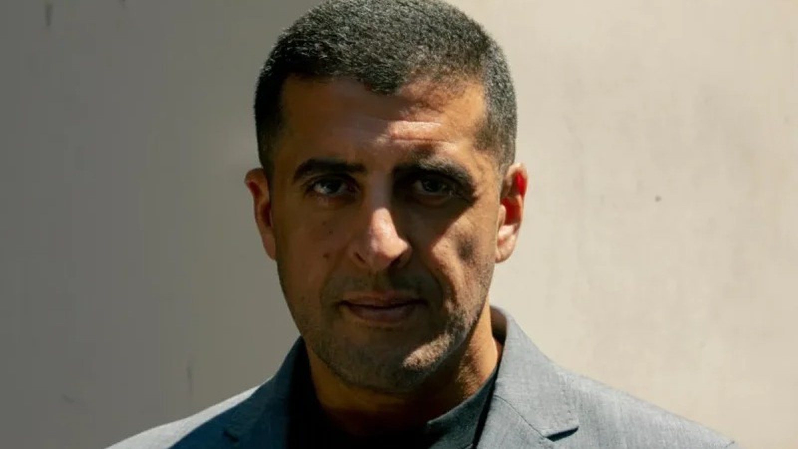 Retrato de homem com cabelo escuro curto e barba rala, usando blazer cinza, olhando à frente