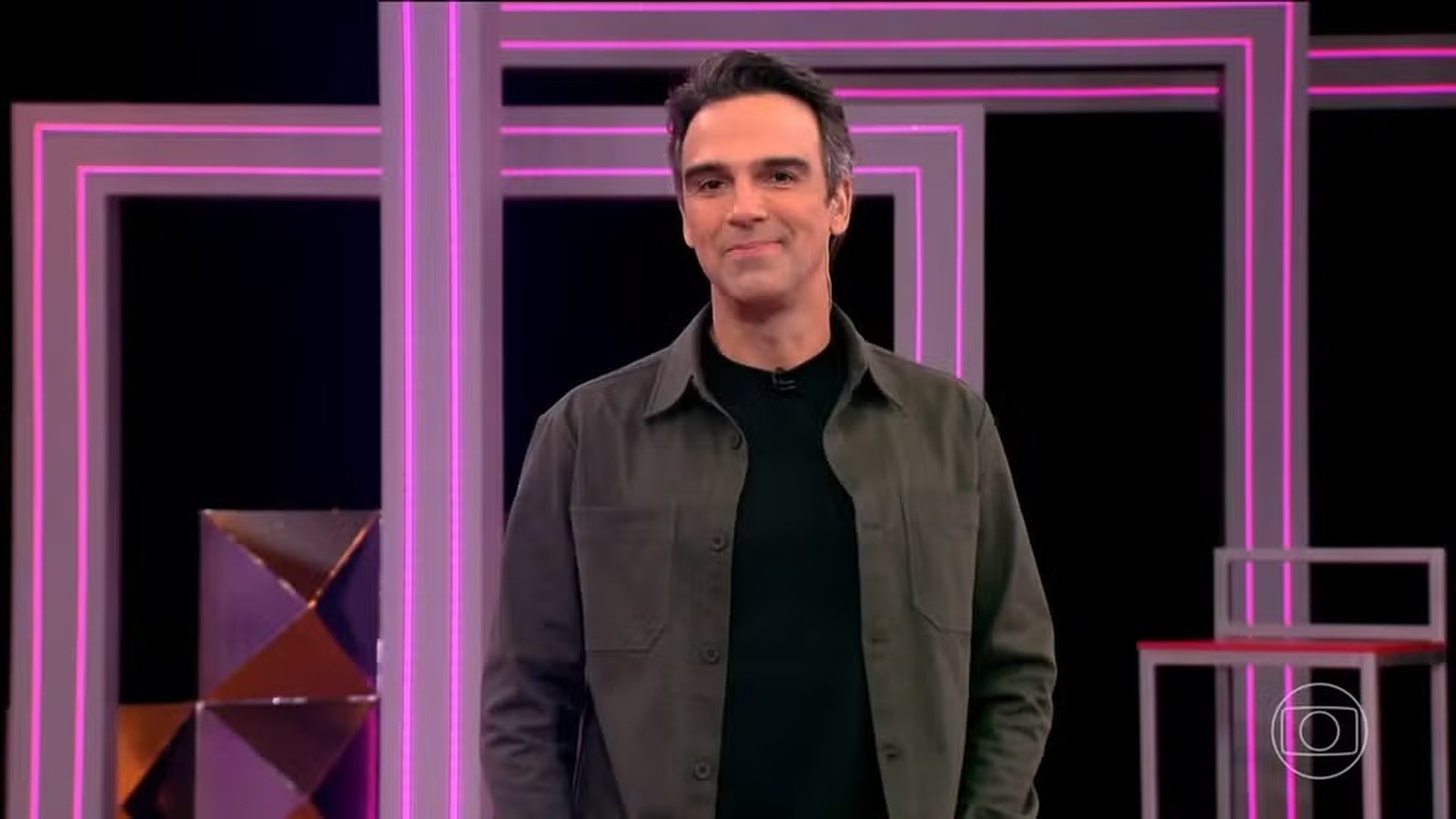 Tadeu Schmidt sorri para a câmera em um estúdio com luzes de neon rosa