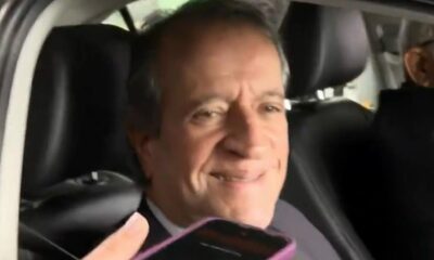 Homem sorrindo dentro de carro, com a mão segurando um celular roxo em primeiro plano