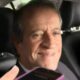 Homem sorrindo dentro de carro, com a mão segurando um celular roxo em primeiro plano
