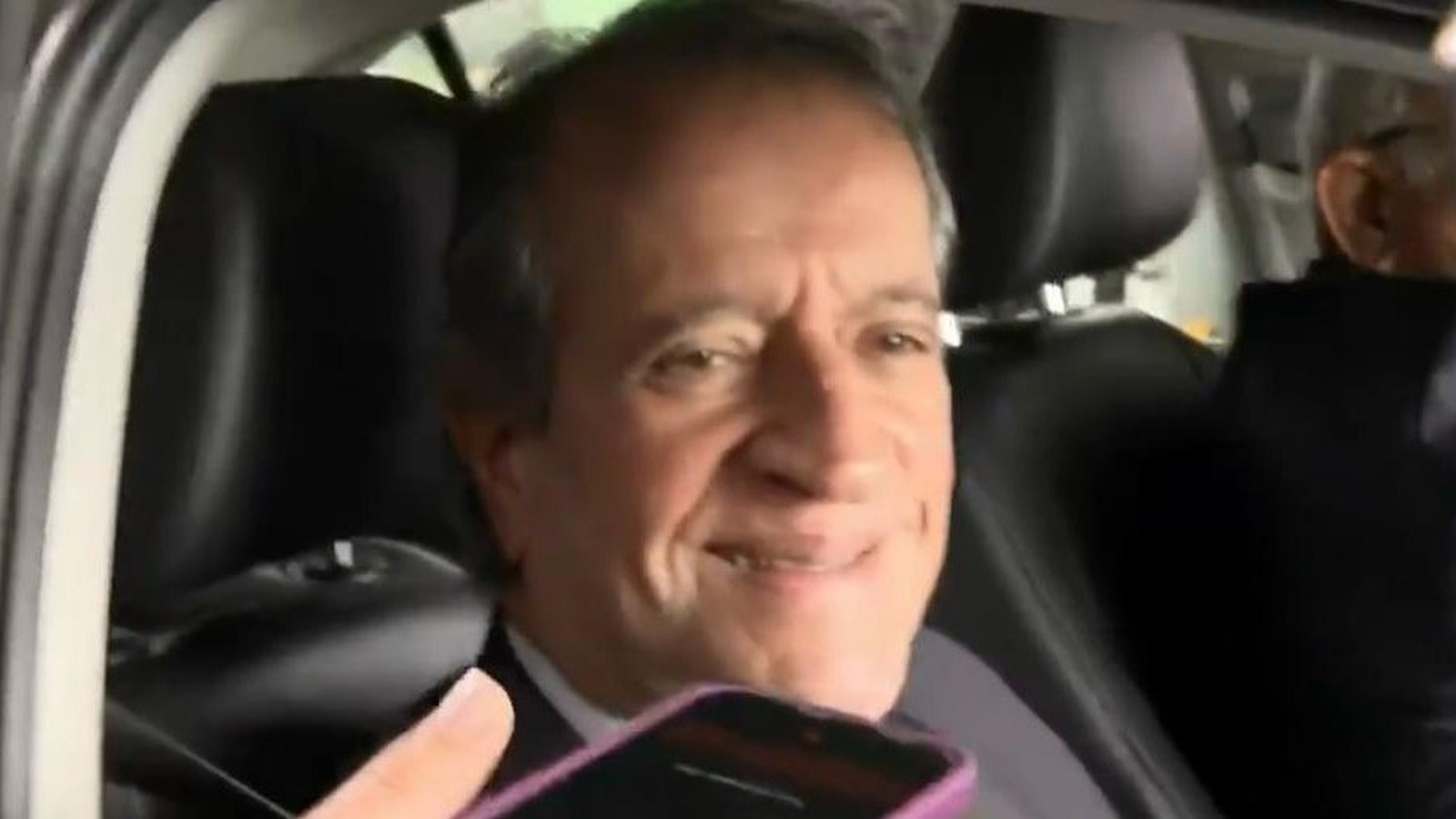 Homem sorrindo dentro de carro, com a mão segurando um celular roxo em primeiro plano