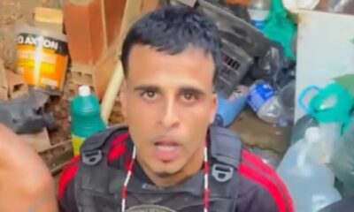 Homem com colete tático, boca aberta, entre entulhos, garrafas e balde Baxton